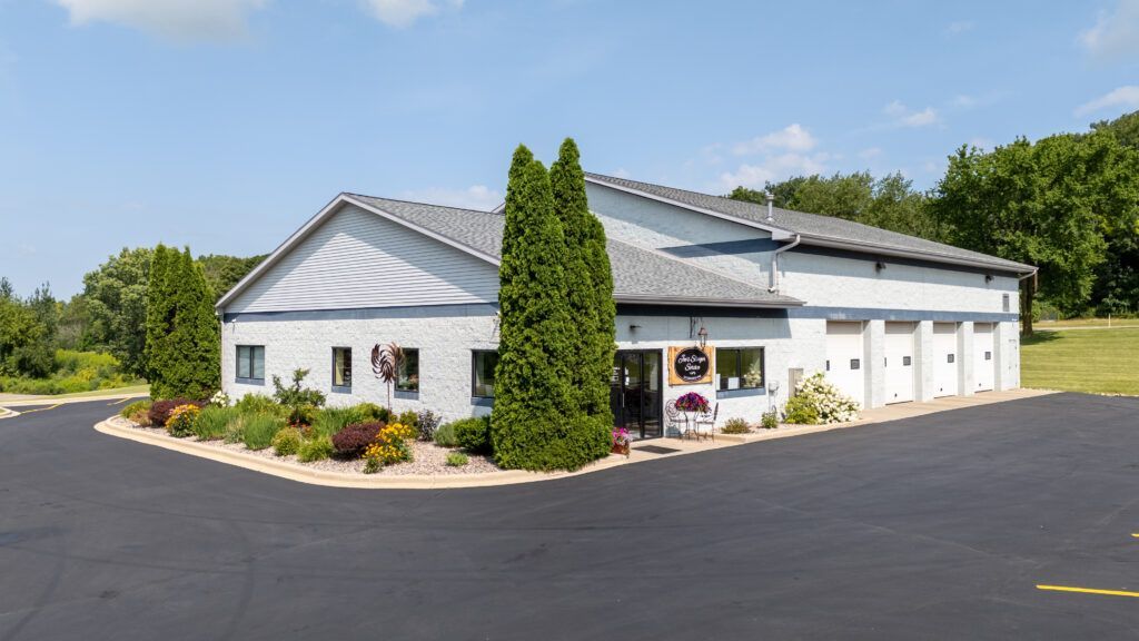 Gray building with a black asphalt driveway, trees, and landscaping.