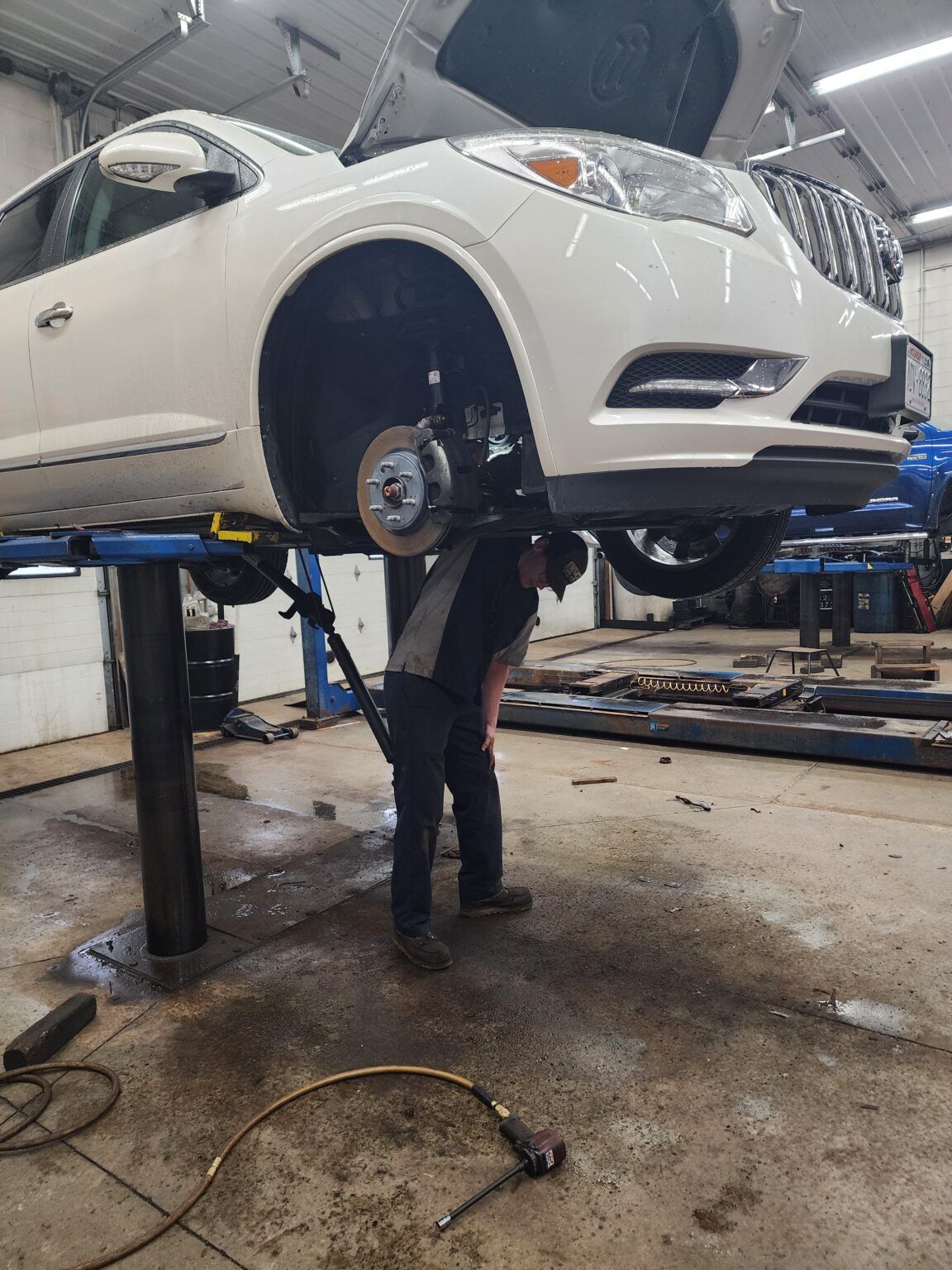 A mechanic working on the front wheel of a white SUV lifted on a car lift in a garage.