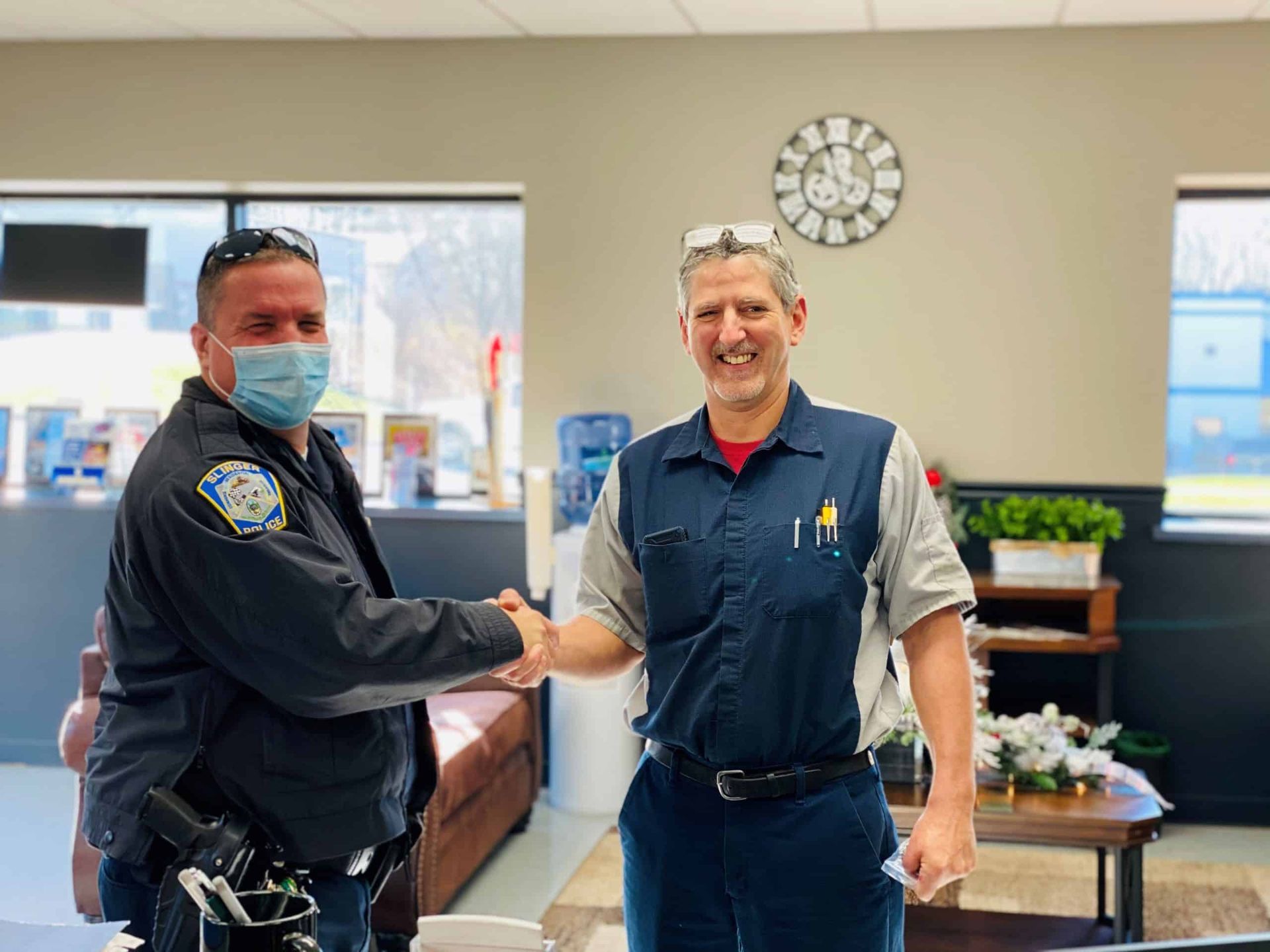 Two men shaking hands in a building. One in a police uniform and mask, the other in mechanic attire.