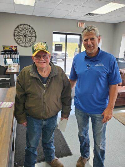 Two men smiling inside a building. One wears a green hat, the other a blue shirt.