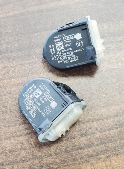 Two black TPMS sensors on a wood surface. Each has a white plastic connector and printed details.