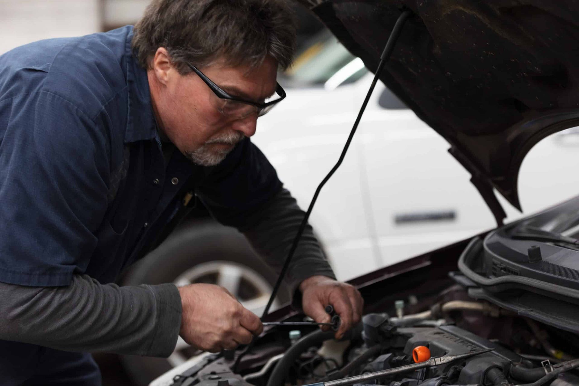 Mechanic in blue work shirt, glasses, working on a car engine in a shop.