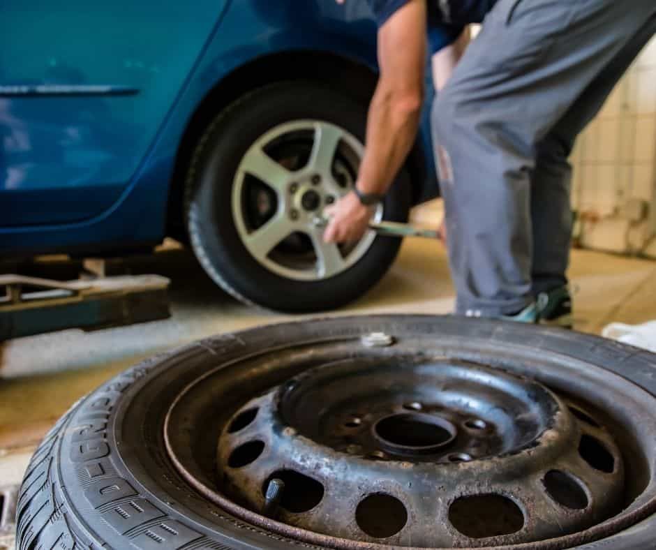 Person changing a car tire with a wrench; spare tire in foreground, blue car in a garage.