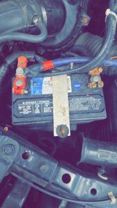 Car battery in engine bay with connected terminals and hold-down bracket.