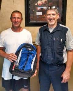 Two men pose with a blue backpack. One man holds it, the other smiles. Indoors, artwork in background.