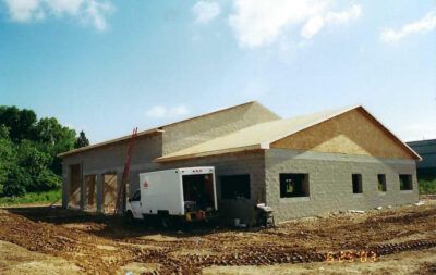 Building under construction, light-colored concrete block exterior, plywood roof, white truck parked at entrance, blue sky.