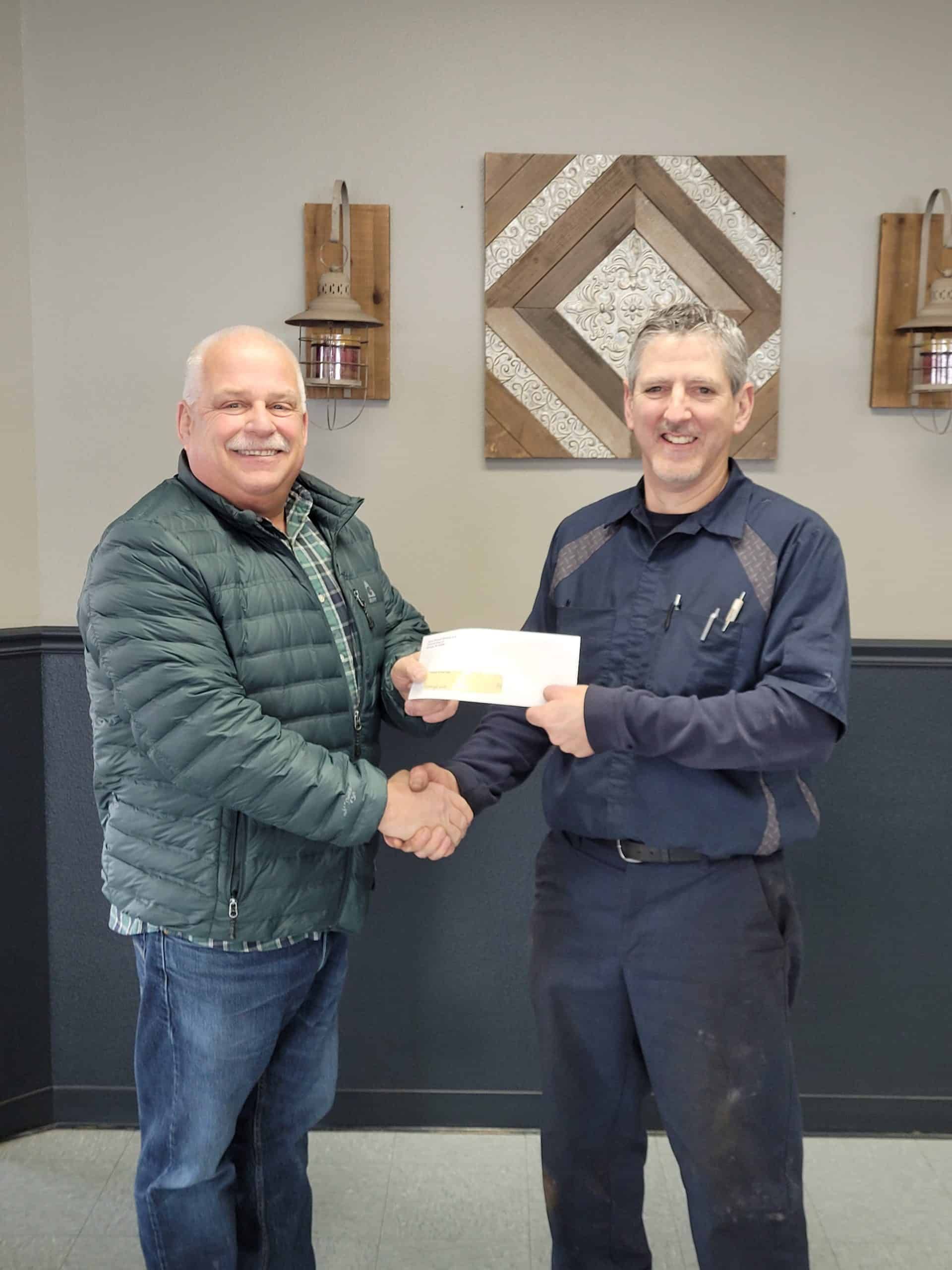 Two men shaking hands, one holding a check. Interior setting with wall art.