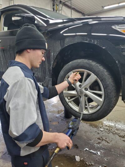 Person inflating a car tire at a service station. The car is black, and the ground is wet.