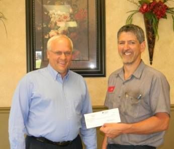 Two men, one in a blue shirt, the other in a work shirt, smiling. One holds a check.