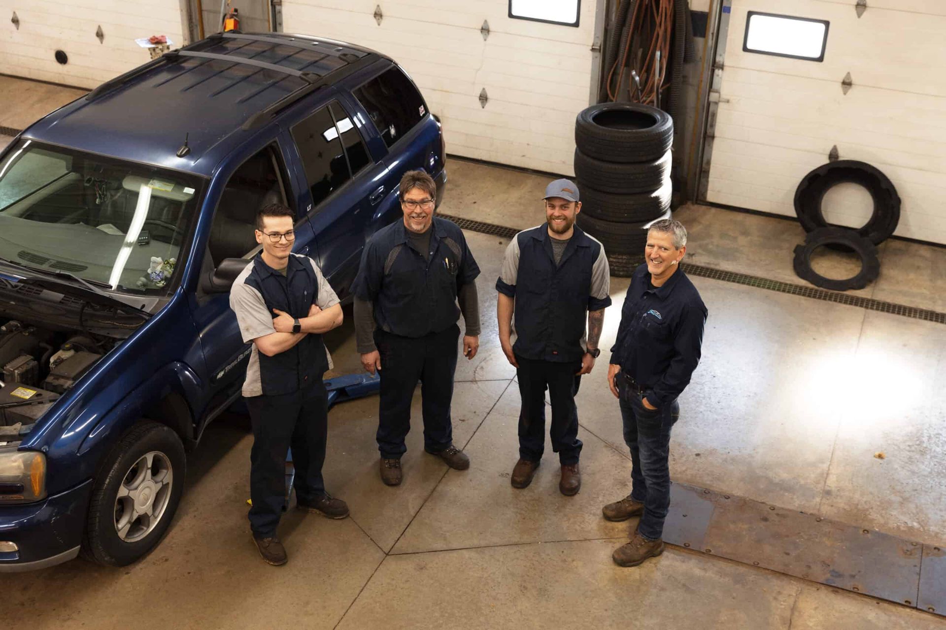 Four mechanics stand near a blue SUV in a garage. One has arms crossed. Tires and tools are visible.