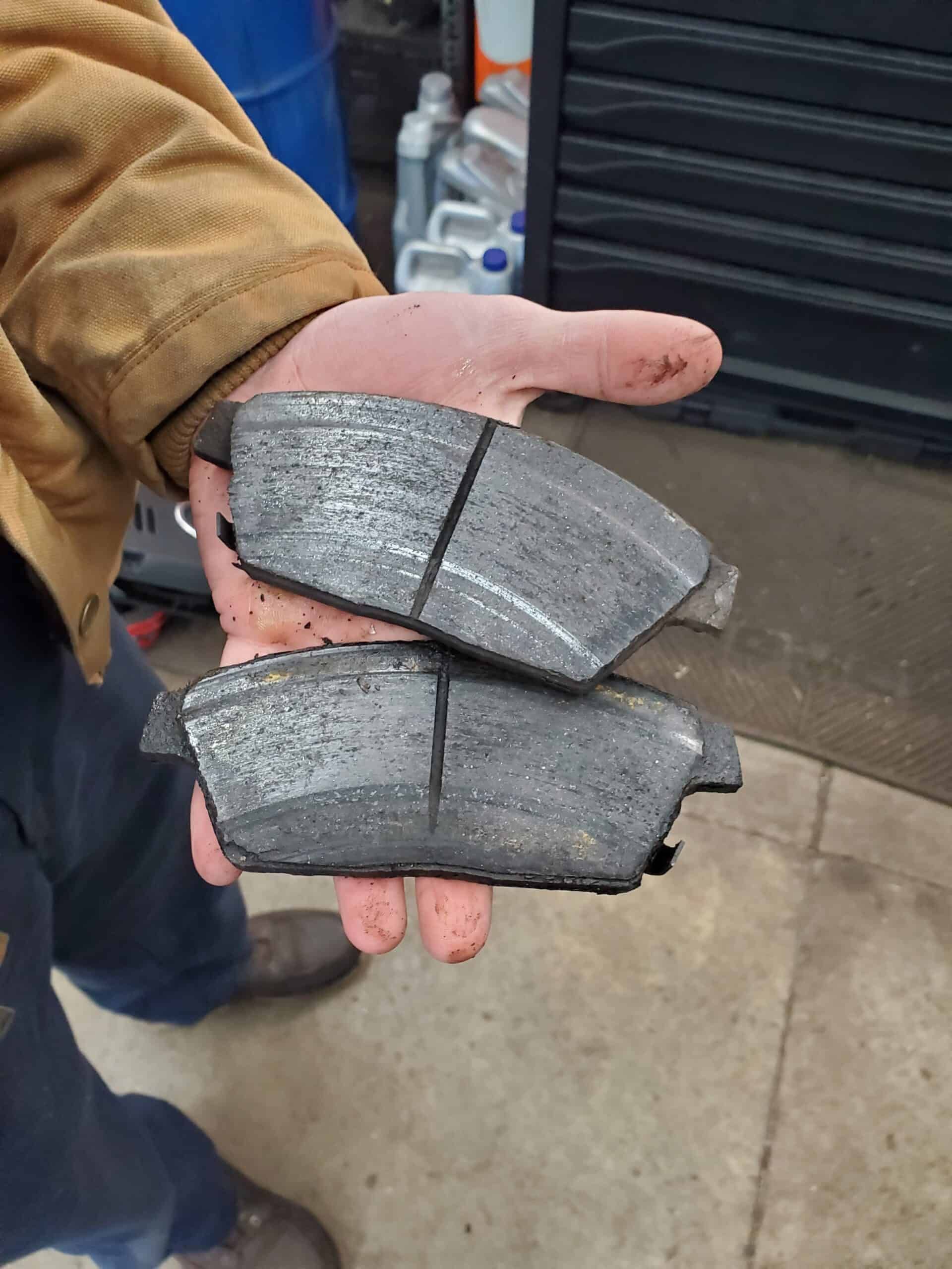 A person holding two worn brake pads, likely from a car, in a garage setting.