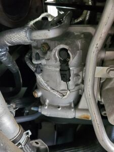 Silver car air conditioning compressor with attached wiring and tubing.