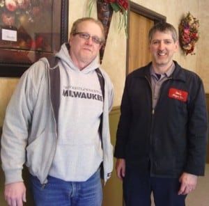 Two men standing inside a building. One wears a grey hoodie and jeans. The other wears a navy jacket with a name tag.