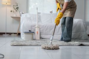Person mopping a tiled floor in a bright bedroom with a gray rug and white bedding