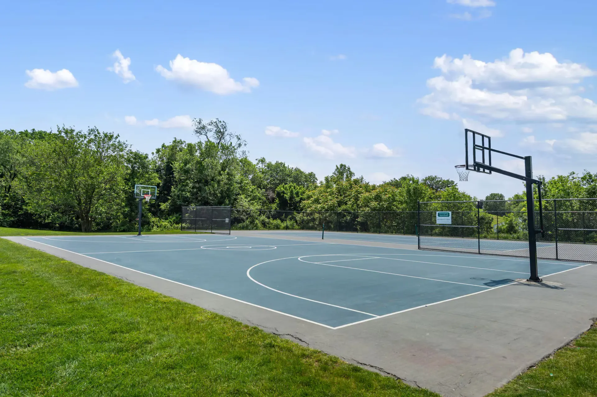 Outdoor basketball court with hoops, surrounded by trees and a chain-link fence.