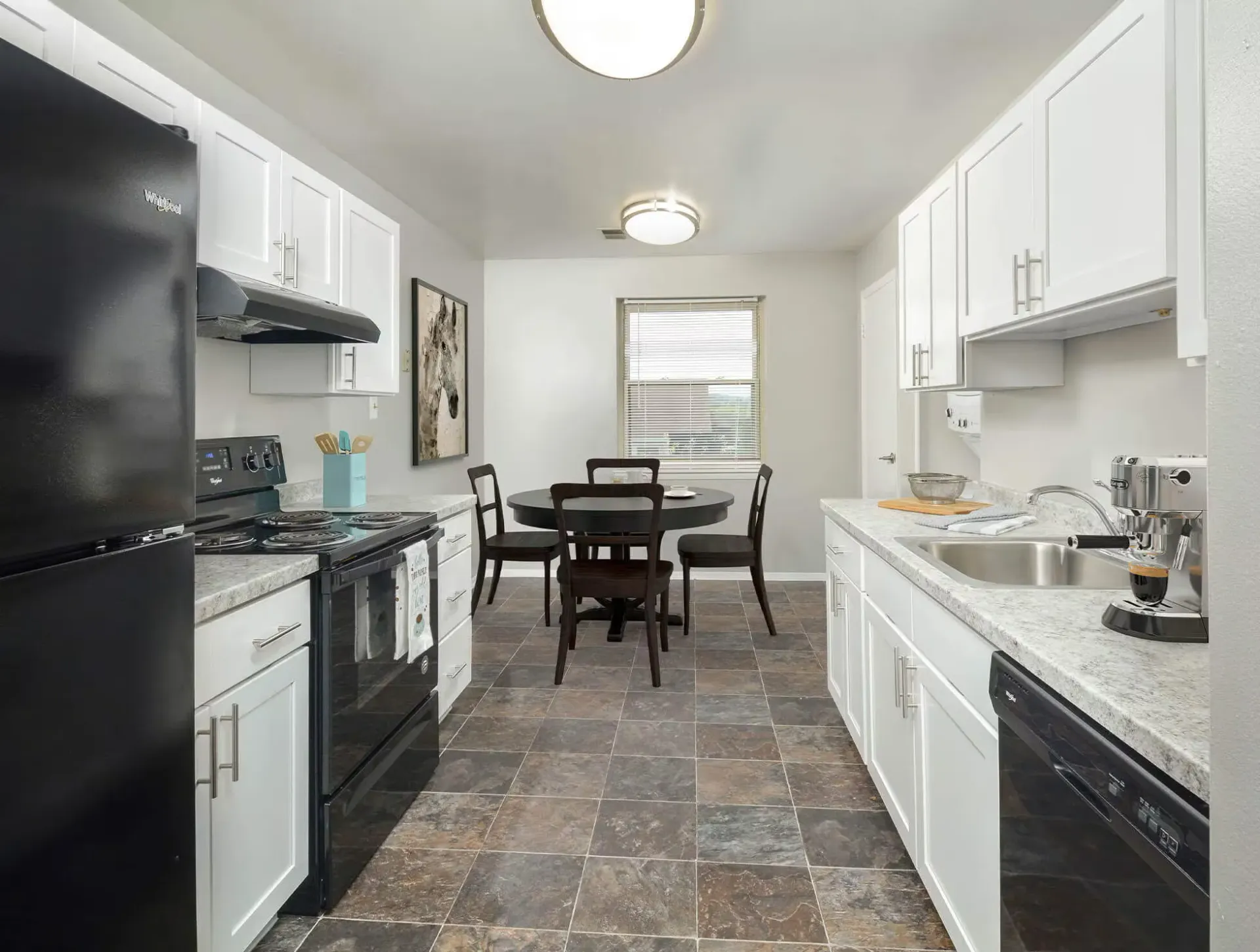 Bright galley kitchen with white cabinets, black appliances, and a round dining table near a window.