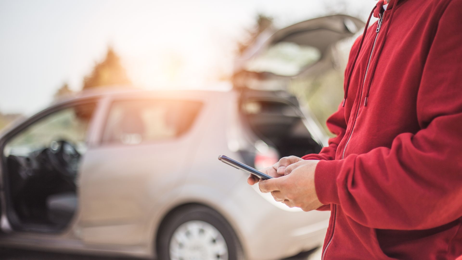 Person in red hoodie using a phone near a car with an open trunk; bright sunlight.