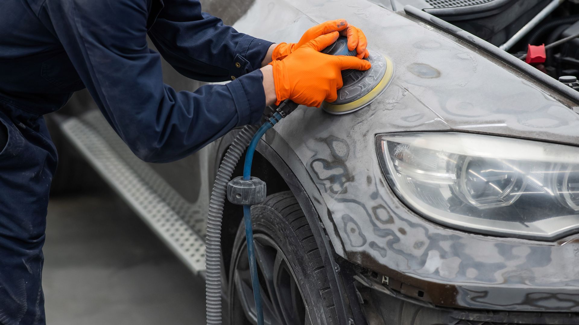 Person sanding a car fender with a power sander, wearing orange gloves, in a shop.