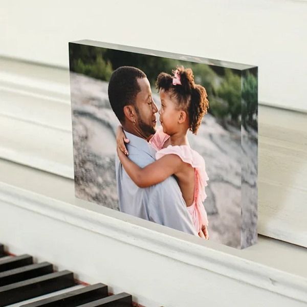 Les mariés sourient en portant un bouquet. Le marié est en costume, la mariée en robe blanche. Le décor est lumineux et ensoleillé.