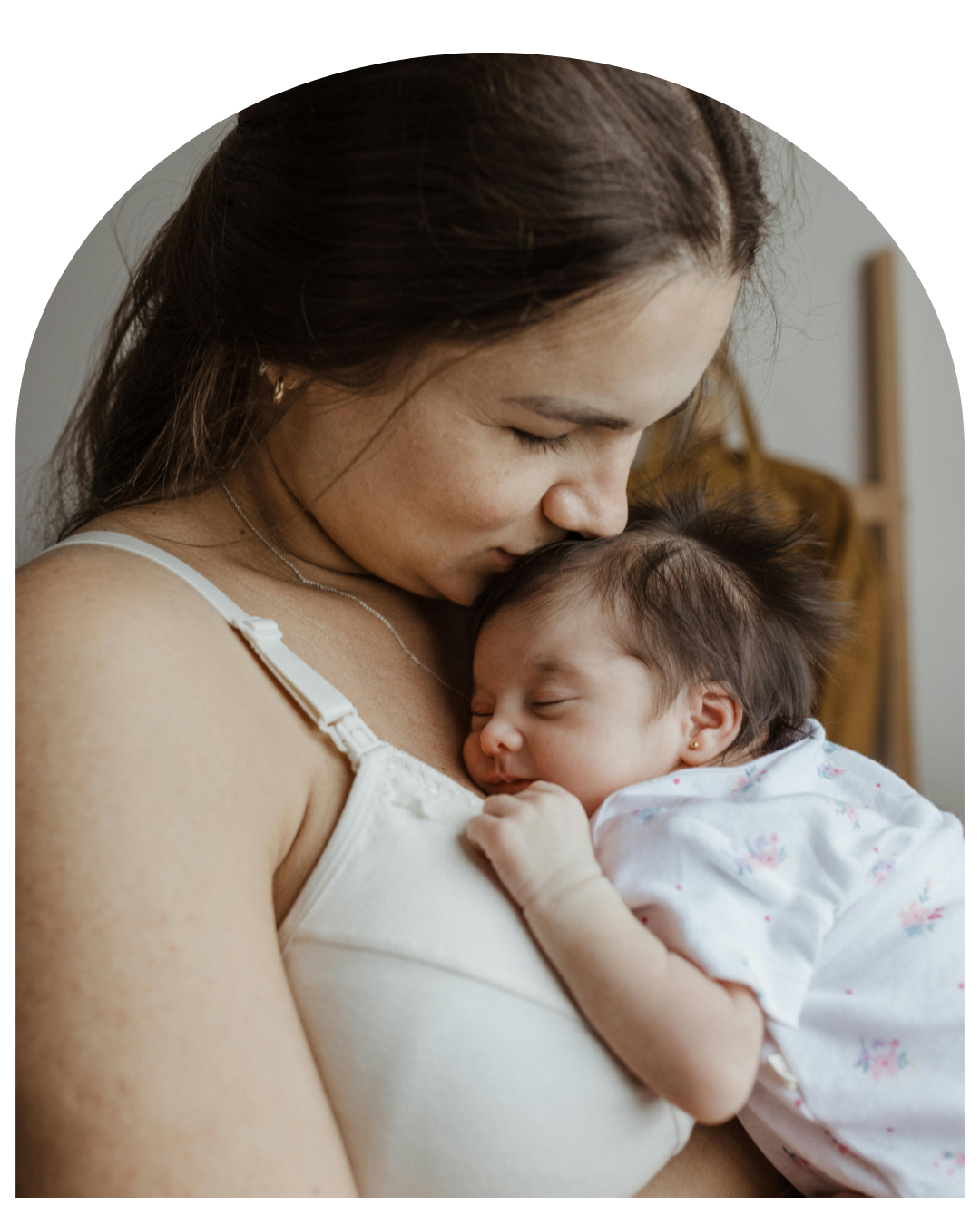 Woman in a white top gently holding and kissing a sleeping baby indoors.