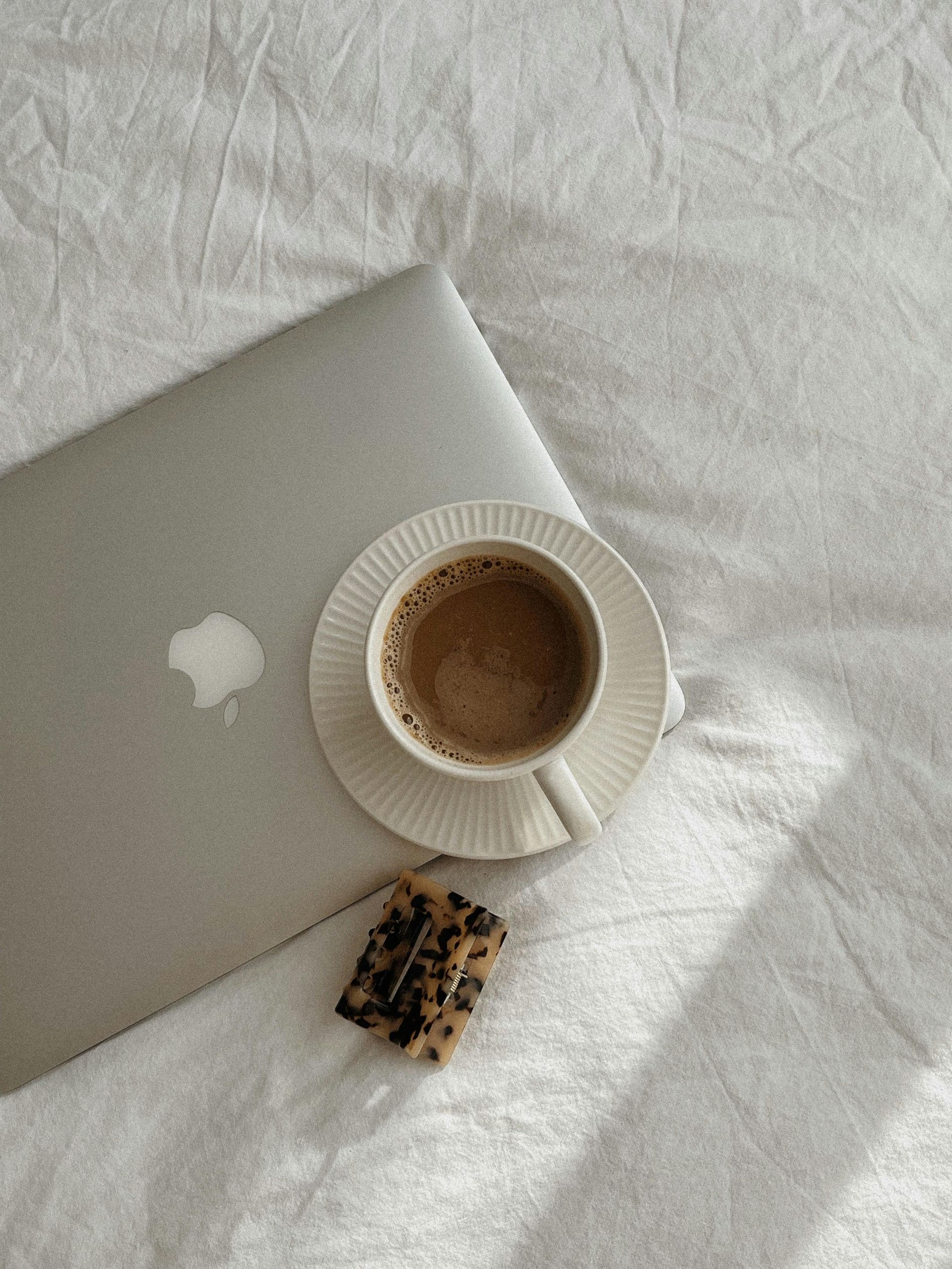 Coffee in a white cup beside a silver laptop on a sunlit white bed, with a cookie nearby.