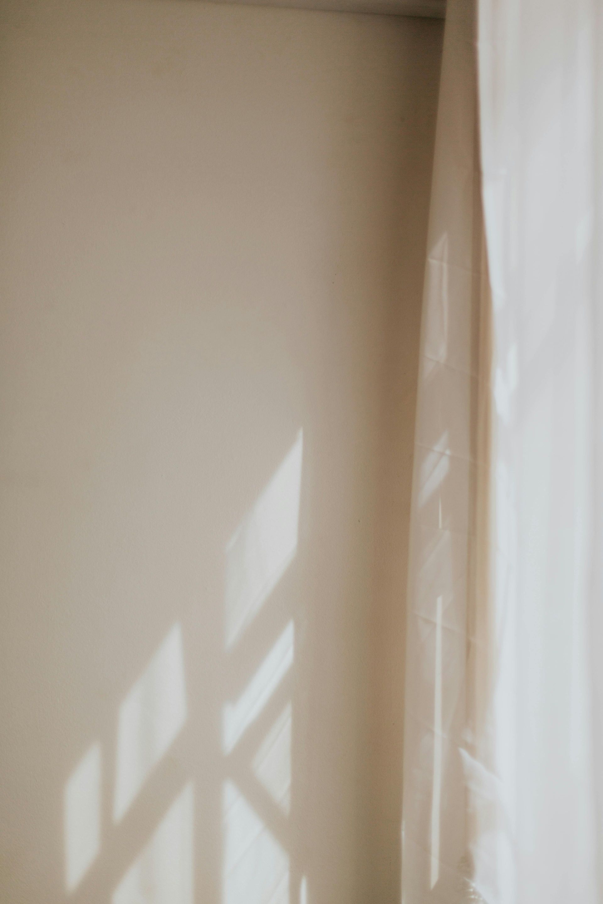 Sunlit beige wall corner with window shadows and sheer curtain