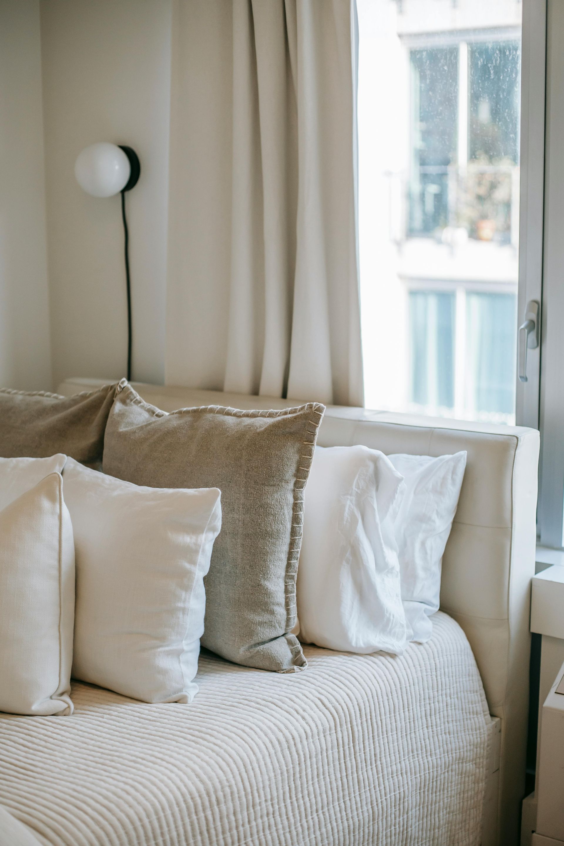 Cozy beige bedroom corner with a bed, textured pillows, and a window with curtains and soft daylight