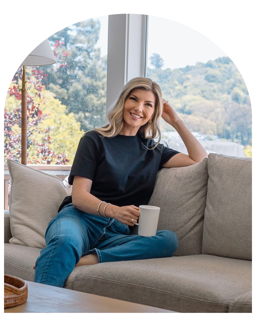 Smiling woman seated on a beige sofa by a window, holding a mug in a bright living room.
