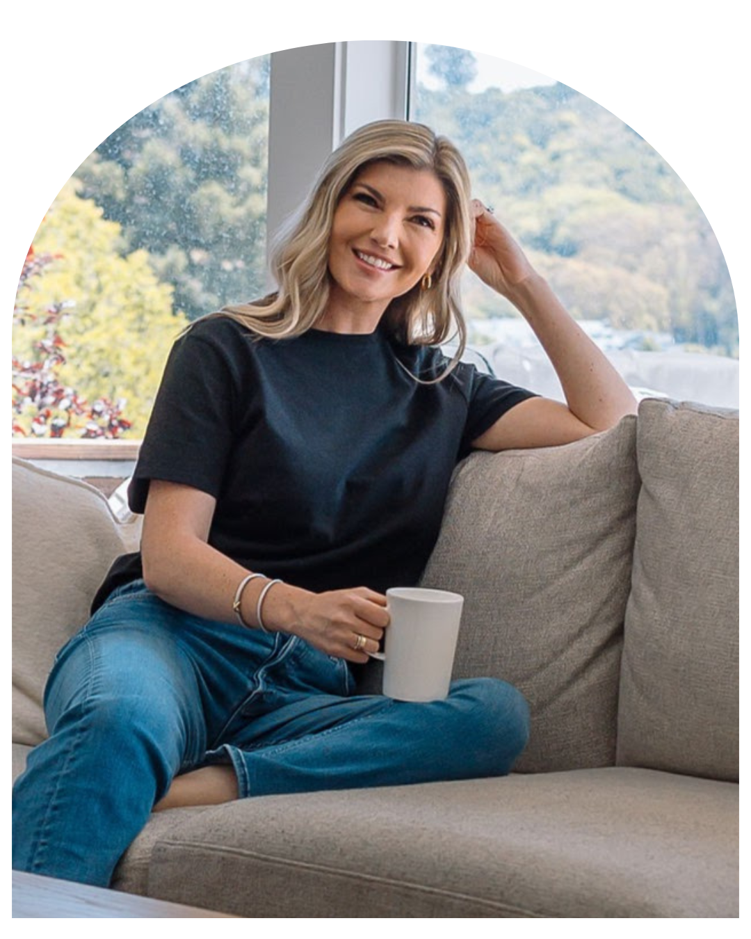 Smiling woman in a black top and jeans sitting on a couch, holding a mug by a window with mountain views