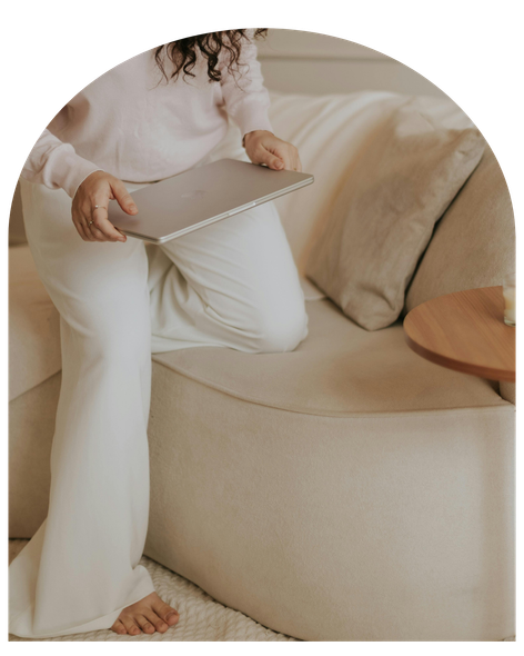 Person in cream clothes holding a laptop while sitting on a beige sofa.