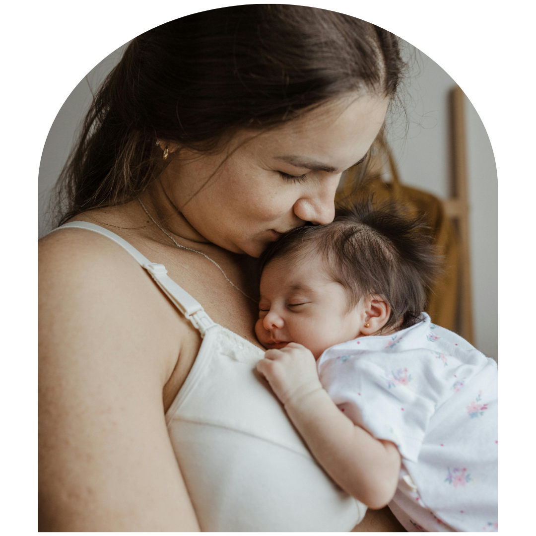 Mother gently kissing sleeping baby in a white crib room