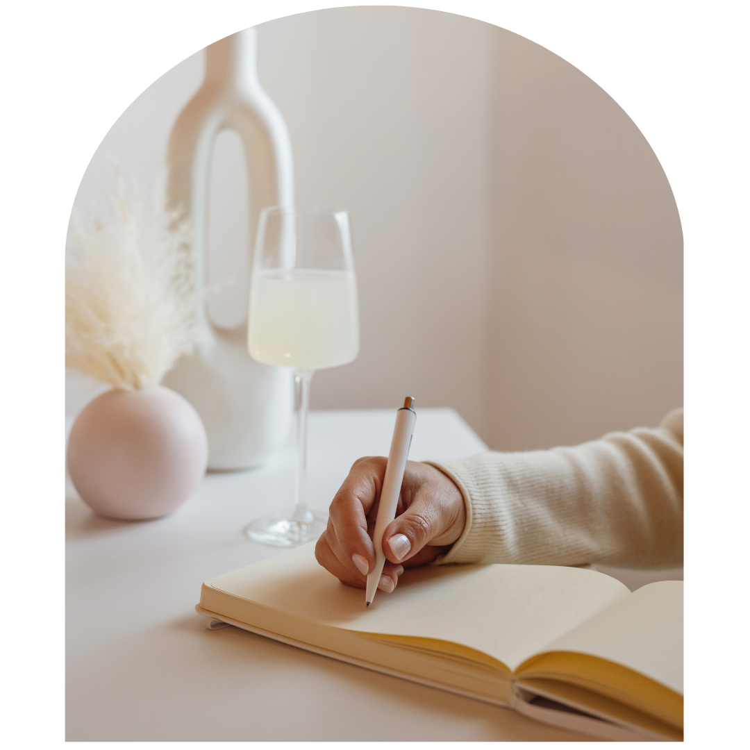 Hand writing in a notebook beside a lit lamp in a soft beige workspace