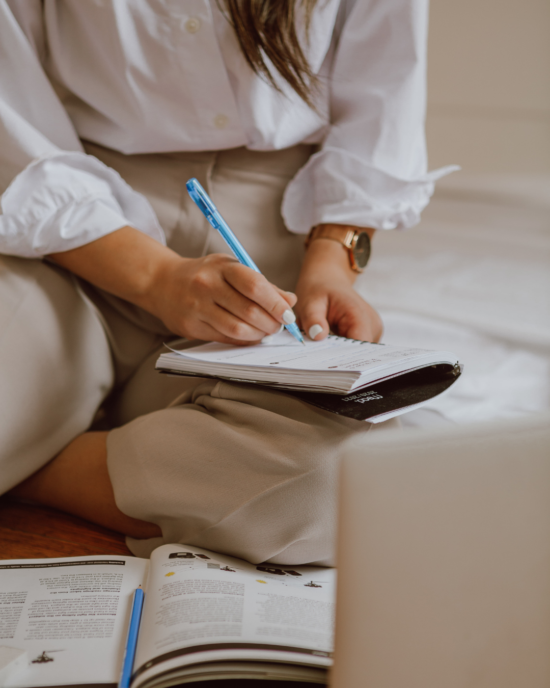 Person writing in a notebook on a sofa, with an open book nearby.