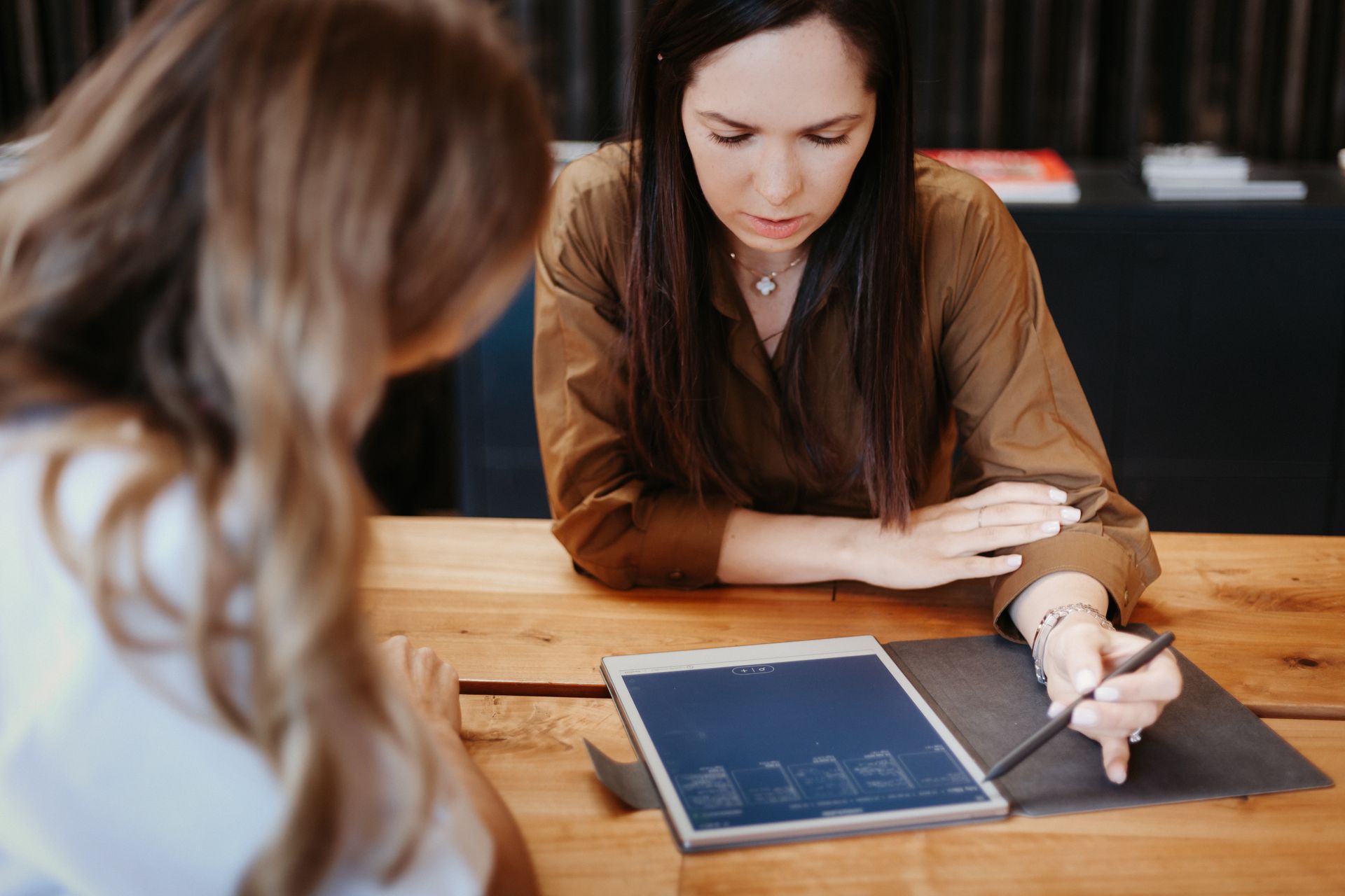 Two women at a wooden table, one points at a tablet with a stylus, the other looks on.