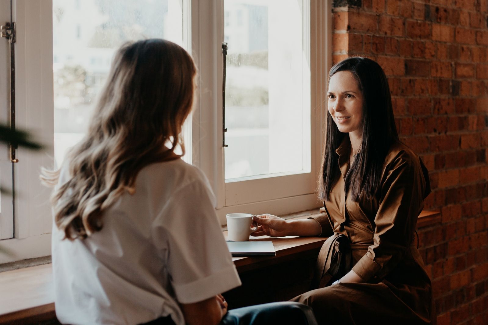Two women talking at a table by a bright window in a cozy café