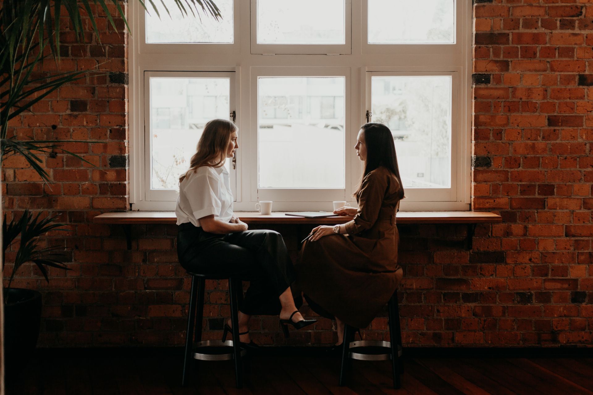 Two people sit at a small table by a bright window in a brick-walled cafe.