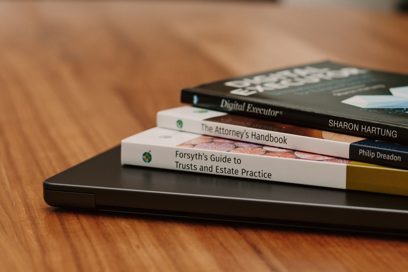 Three stacked books resting on a laptop on a wooden table, with a blurred book in the background