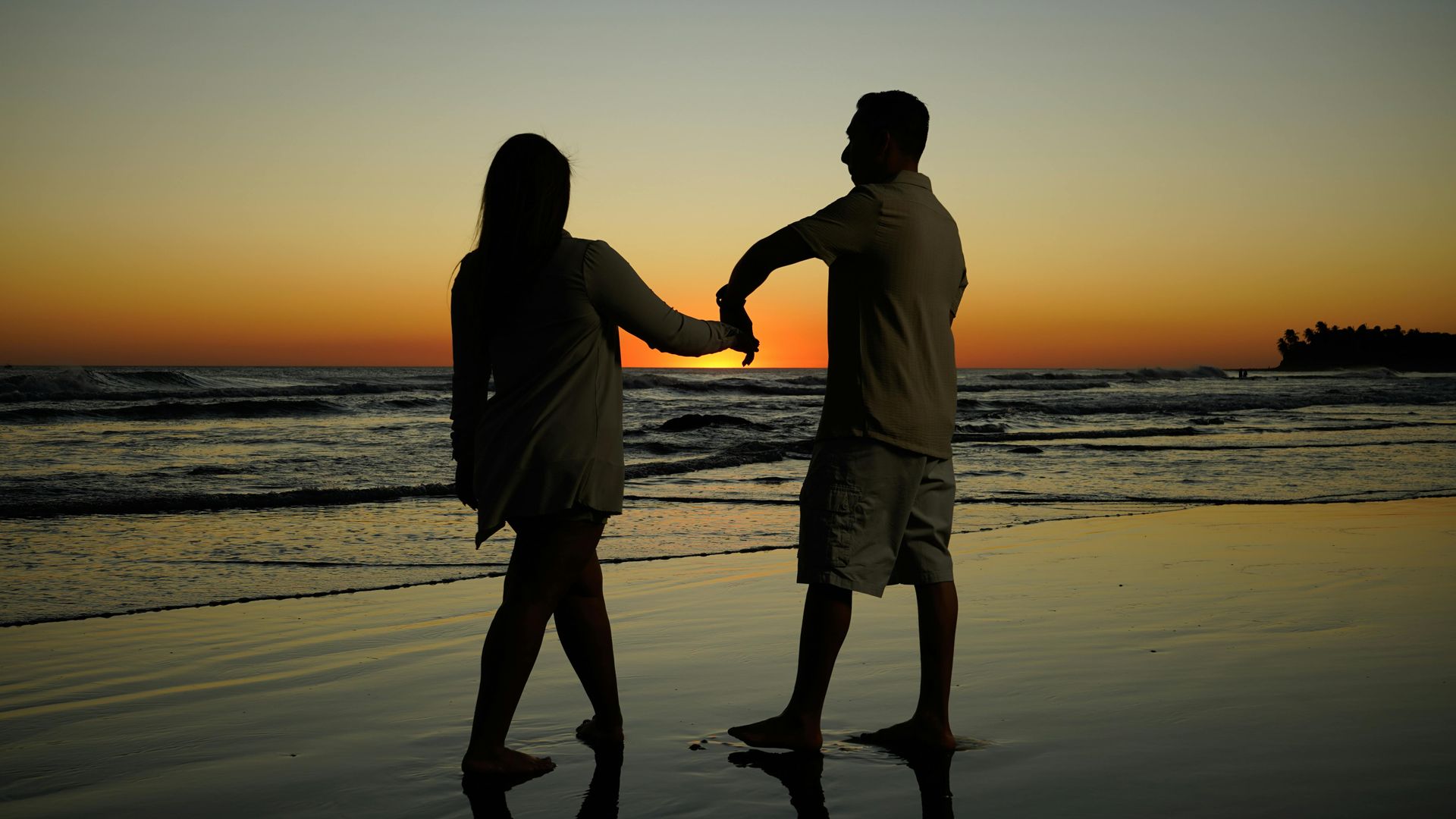 Couple holding hands, silhouetted on beach at sunset.