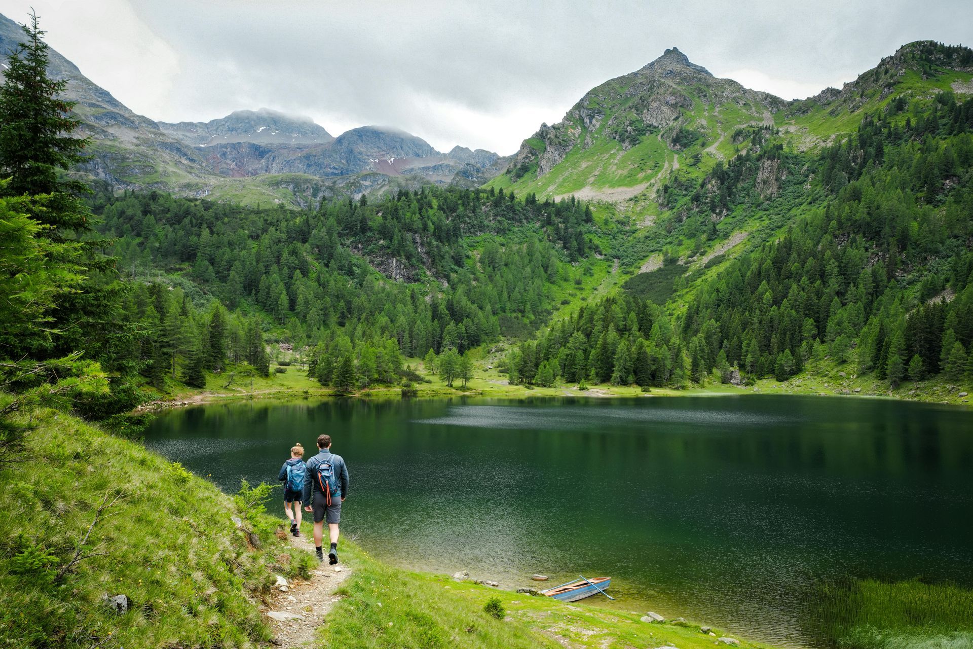 Hikers on a lakeside trail beside a clear mountain lake and green forested peaks