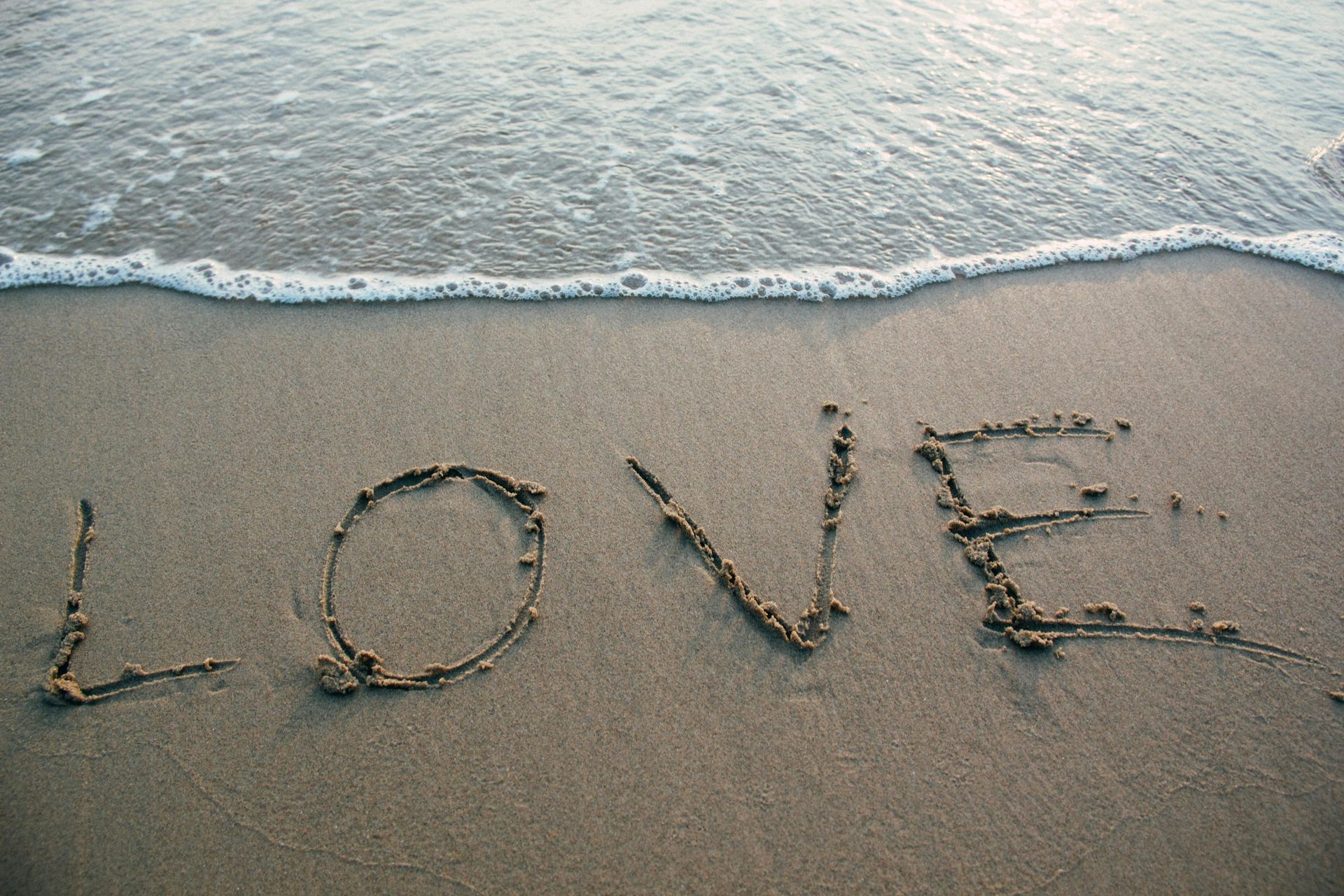 Word “LOVE” written in sand on a beach beside gentle ocean waves.