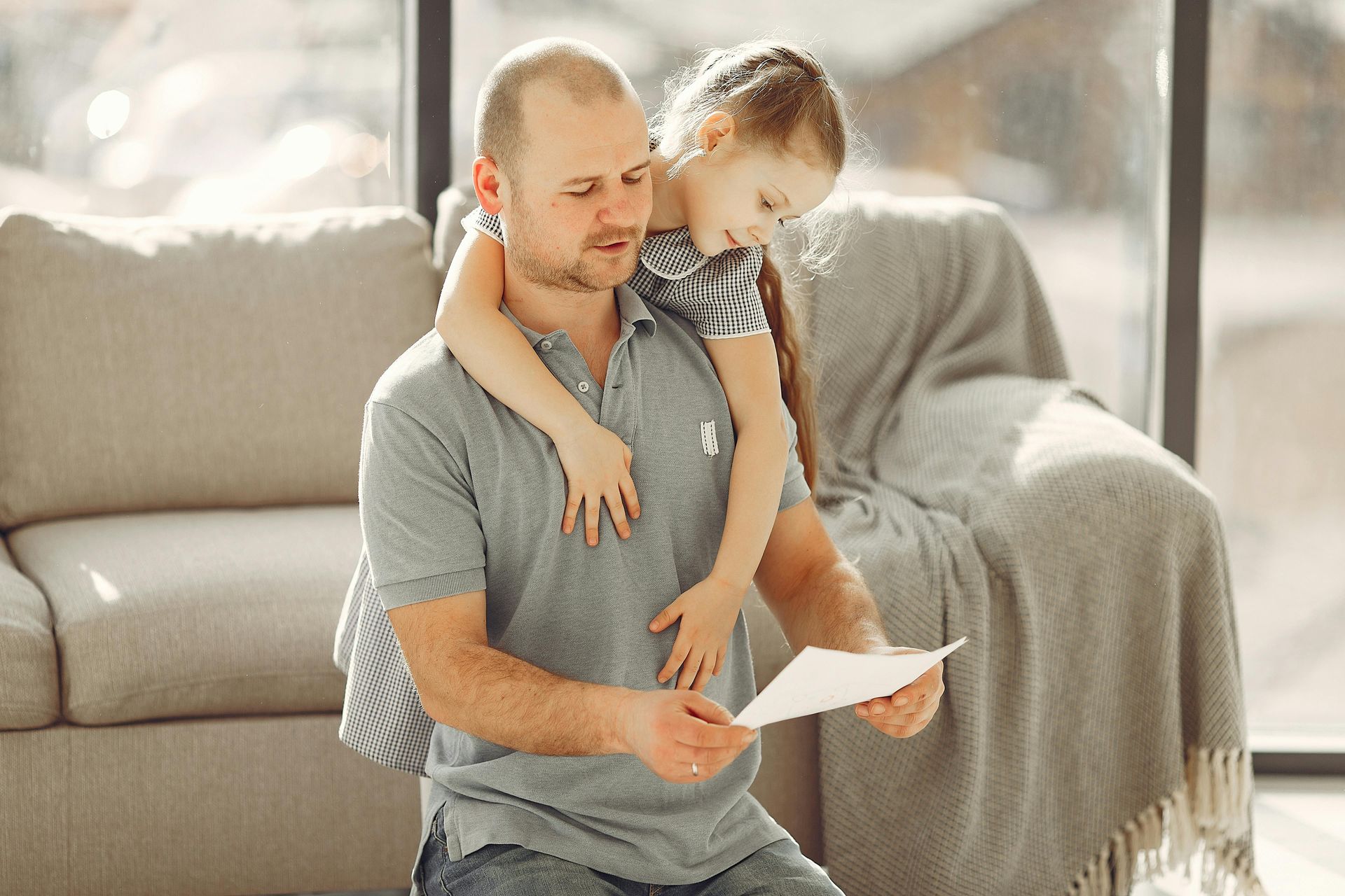 Adult and child sitting on a couch, reading a paper together in a bright living room.