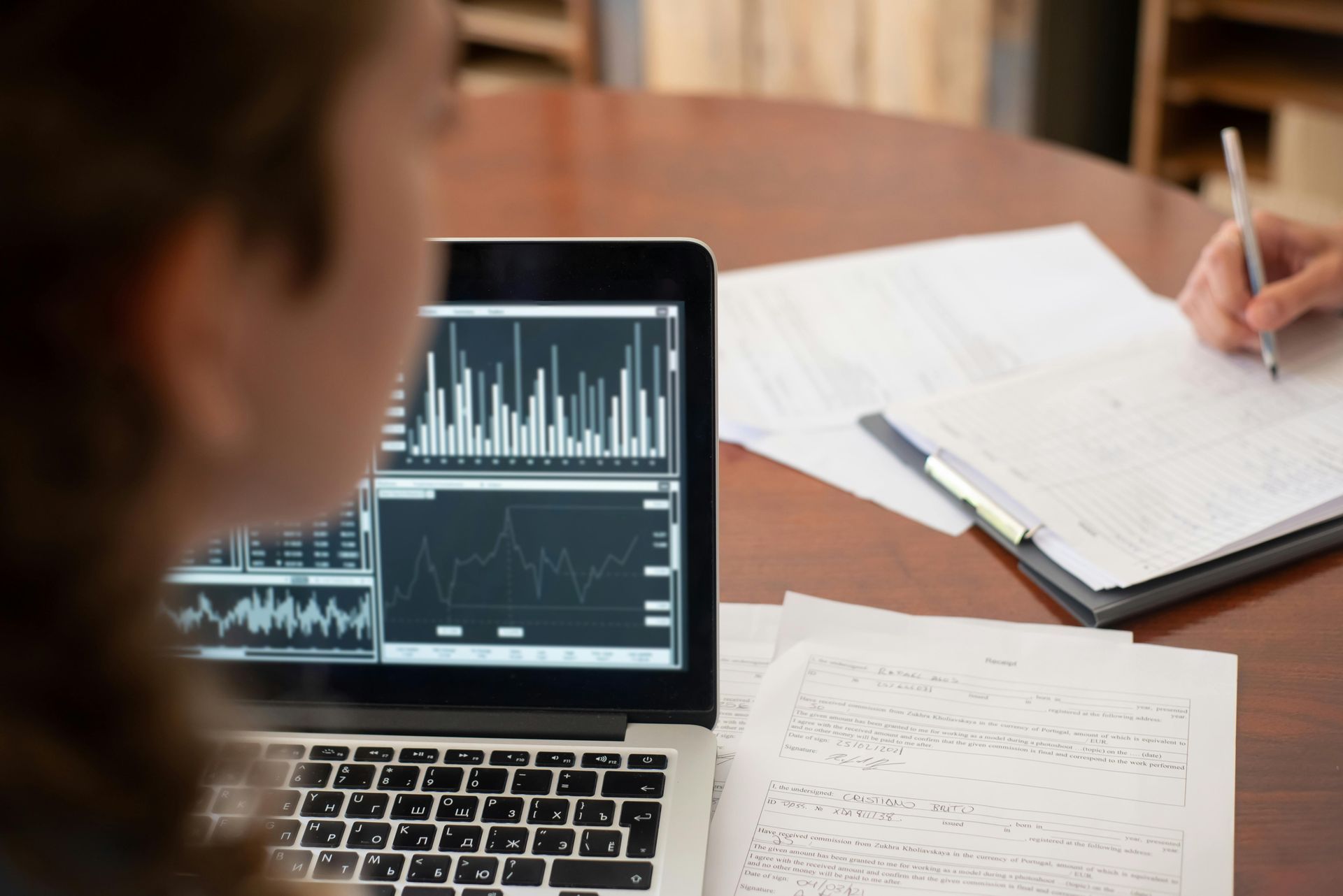 Laptop with charts on a desk beside paperwork and a hand writing notes in a notebook