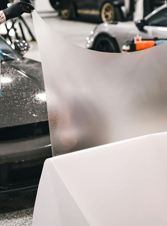 A gloved hand holds a large, translucent matte film over a black car in a garage workshop.
