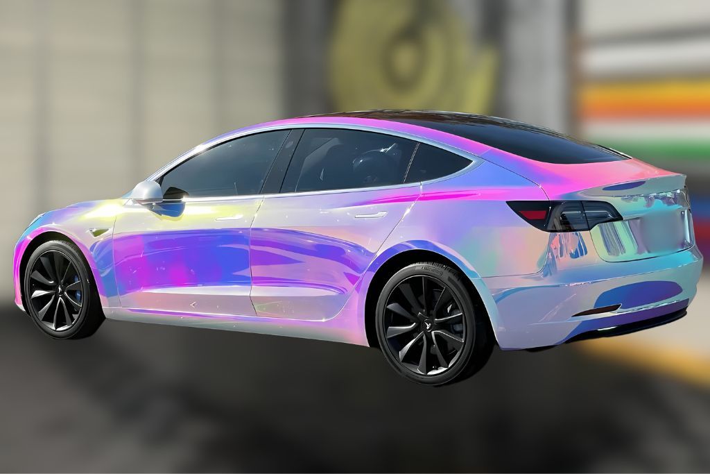 A Tesla Model 3 wrapped in a vibrant chrome iridescent finish, reflecting a spectrum of colors including pink, purple, and gold. The dynamic wrap adds a futuristic and eye-catching flair to the car's sleek design.