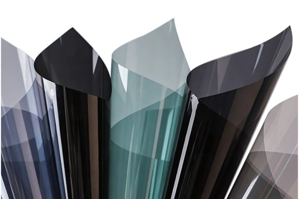 A selection of rolled window tint films in various shades, ranging from dark black to lighter gray and green hues. The films display a matte, non-reflective finish.