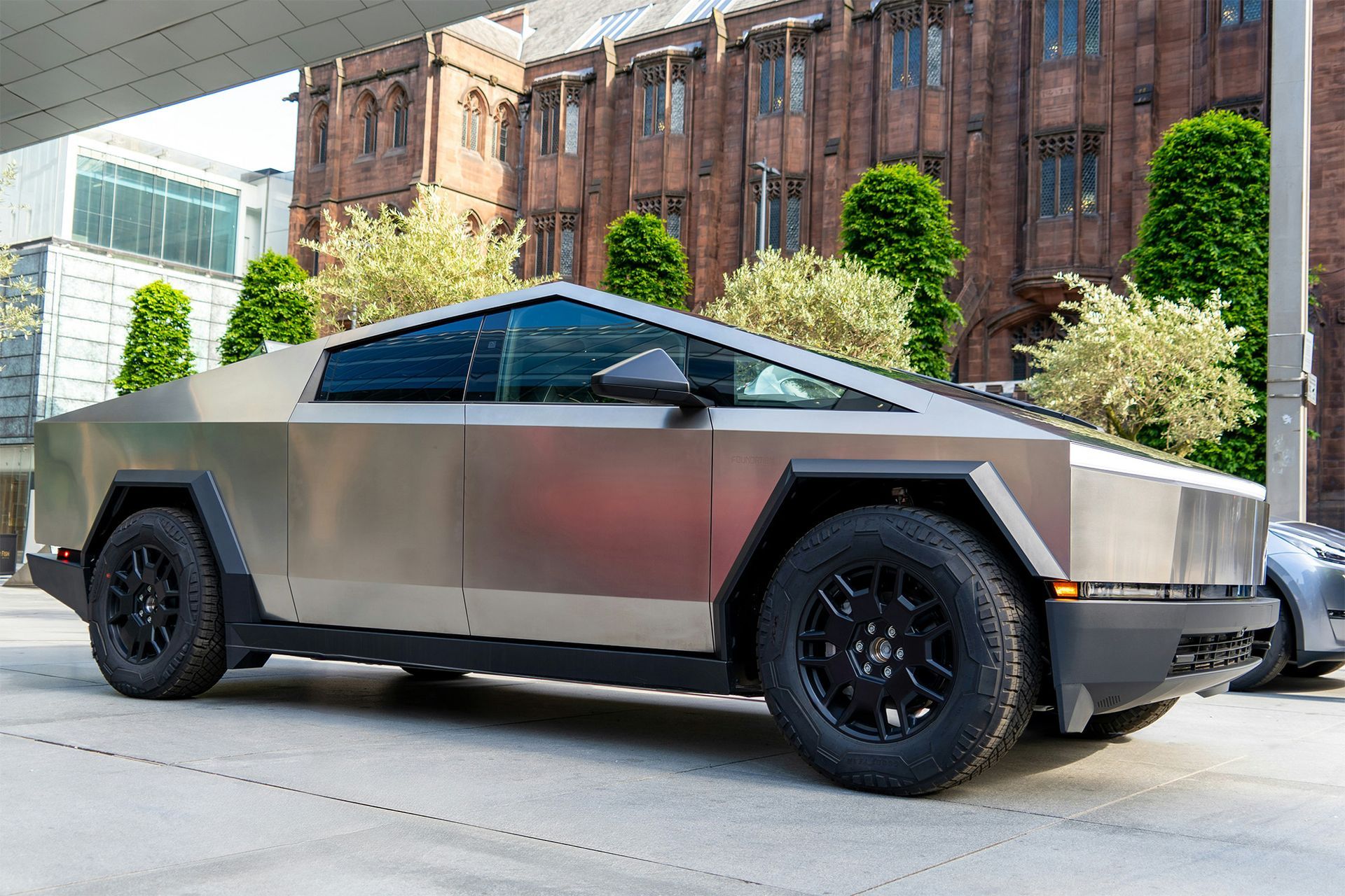 Silver Tesla Cybertruck parked on a city street, near a brick building and greenery.