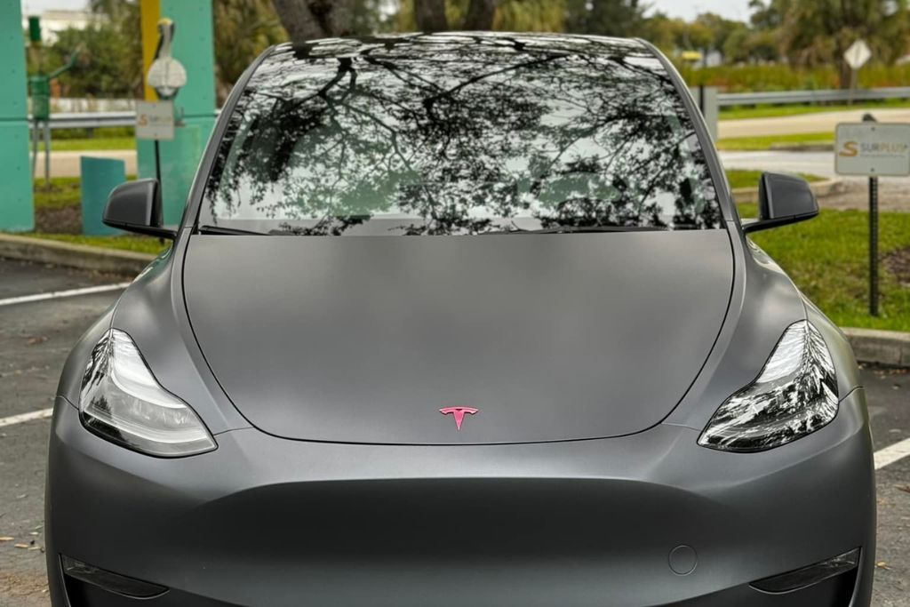 A Tesla Model Y featuring a matte gray vinyl wrap, highlighting its understated and modern look. The matte finish minimizes glare while accentuating the car's clean, futuristic lines, with a pink Tesla logo adding a pop of color.