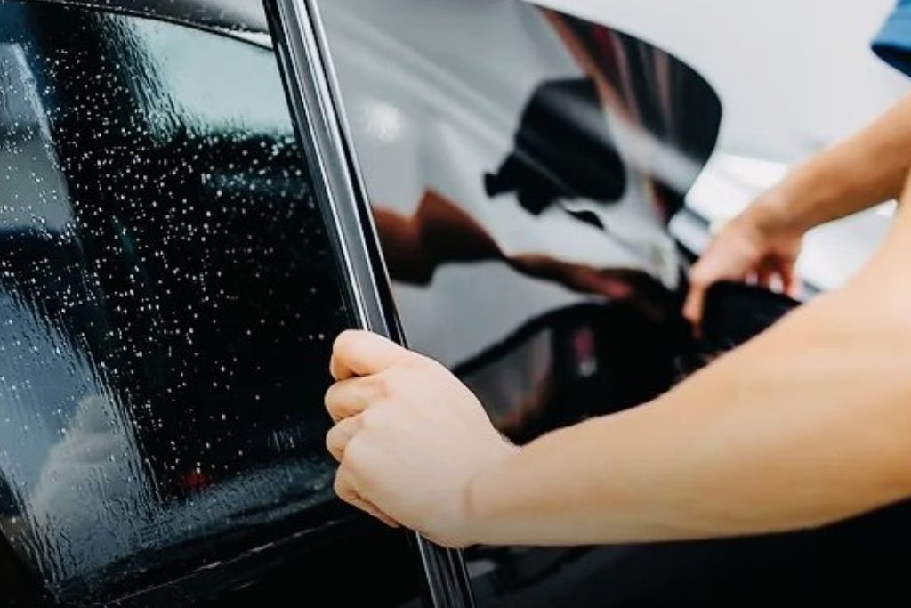 A person carefully applies a tinted film to a car window, smoothing it out over the wet glass surface. The reflection on the window highlights the dark, sleek finish of the tint.
