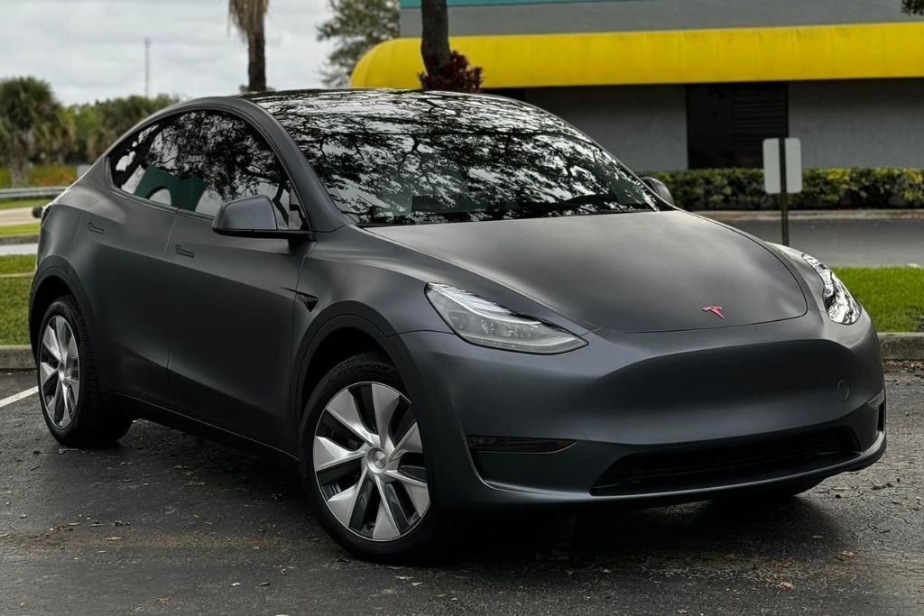 A Tesla Model Y wrapped in a satin gray finish, blending the soft sheen of gloss with the understated elegance of matte. The wrap highlights the car's smooth curves and futuristic design, with a pink Tesla logo adding a modern touch.
