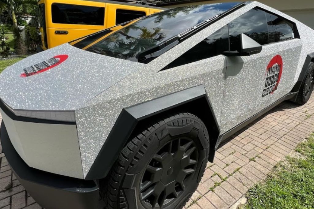A Tesla Cybertruck with a specialty glitter vinyl wrap, showcasing a sparkling silver texture that catches the sunlight. The wrap features a bold red and black circular logo on the side for added personalization.